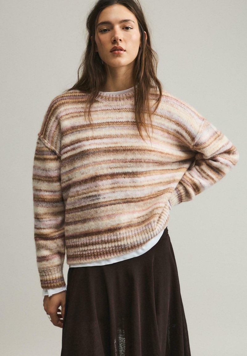 Femme aux longs cheveux bruns portant un pull doux rayé aux tons beige et rose, et une jupe foncée, debout avec une main sur la hanche.