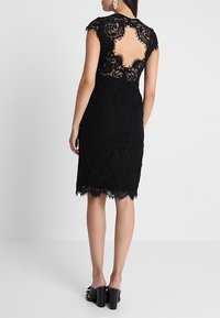 Schwarzes Spitzenkleid mit überschnittenen Ärmeln, offenem Rücken, wellenförmigem Saum und filigranen floralen Mustern, kombiniert mit Sandalen mit klobigem Absatz.