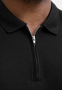 Sort polo shirt med en tekstureret, vaffel-lignende mønster; har en lynlåskrave og kontrasterende sølv lynlåshardware.