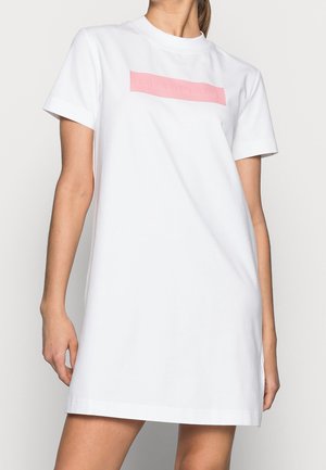 Žena nosící krátké bílé tričko-šaty s růžovým obdélníkovým logem na hrudi.