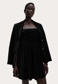 Černé sametové šaty s přiléhavým korzetem a plisovanou sukní, s hranatým výstřihem zdobeným květinovými výšivkami. Nosené pod černým blejzrem.