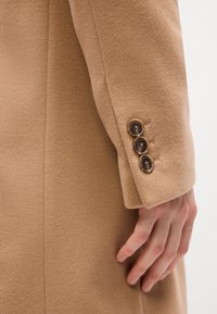 Light beige wool coat with a smooth texture, featuring three dark brown buttons on the cuff and tailored sleeve design.