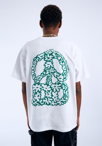 Bílý krátký rukáv tričko s velkým zeleným květinovým symbolem míru na zadní straně, obklopeným textem. Standardní střih.