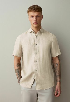 Ung mand med kort hår iført beige kortærmet skjorte og lyse bukser, stående op ad en ensfarvet lys væg.