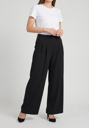 Femme portant une chemise blanche à manches courtes, un pantalon noir taille haute à jambes larges et des chaussures noires à talons hauts, debout devant un fond uni.