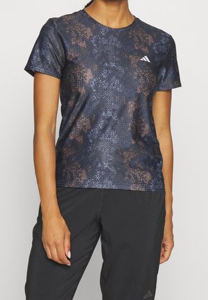Person iført en mørk mønstret Adidas sportstrøje og sorte bukser, stående mod en ensfarvet lys baggrund. Overkroppen er synlig, armene ned langs siden.