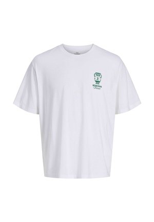 Nastolatek z kręconymi włosami, ubrany w białą koszulkę z zielonym logo, dżinsowe spodenki, białe skarpetki i beżowe buty, stoi i uśmiecha się, zwrócony w lewo.