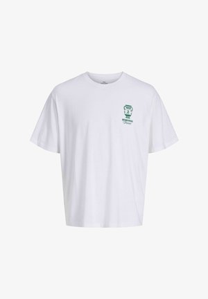 Dospievajúci chlapec s kučeravými vlasmi, oblečený v biele tričko so zeleným logom, rifľové krátke nohavice, biele ponožky a béžové topánky, stojaci a usmievajúci sa, otočený smerom doľava.