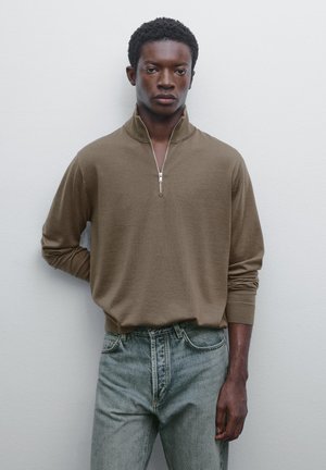 Ung man som står mot en vit vägg, iförd en brun halvdragkedjeströja och ljusblå jeans, med en hand bakom ryggen.