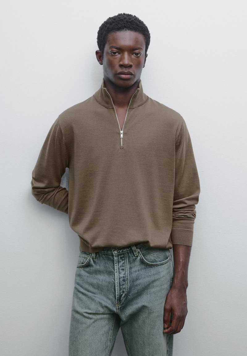Young man standing against white wall, wearing a brown half-zip sweater and light blue jeans, with one hand behind his back.