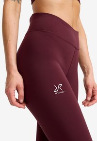 Närbild av en person som bär högmidjade bordeauxfärgade leggings med en vit RVRC-logga på låret mot en enfärgad bakgrund.