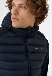 Młody mężczyzna z kręconymi włosami, noszący granatową, ocieplaną kamizelkę Champion z zamkiem błyskawicznym i kapturem, patrzy w bok.