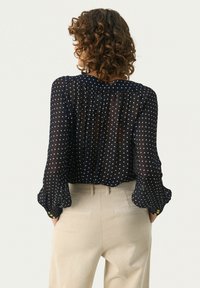 Blouse marine à pois avec un tissu transparent, des manches longues et un dos froncé. Associée à un pantalon en velours côtelé beige présentant des rayures verticales.