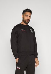 Fekete pulóver, melyen hímzett logók találhatók a mellkasrészen és az ujjon, párosítva ugyancsak fekete melegítőnadrággal, ami laza szabást és egyszerű dizájnt mutat.