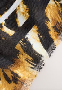 Schwarz und senfgelb gemusteter Stoff mit ausgefransten Kanten, gewebter Textur und abstraktem Pinselstrich-Design vor hellem Hintergrund.