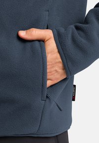 Giacca in pile blu navy con tasca con zip, superficie texturizzata e etichetta Polartec sul lato. Tessuto morbido e vestibilità rilassata.