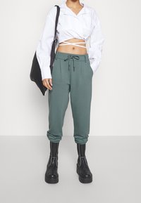 Avskuren vit långärmad skjorta, gröna joggingsbyxor med dragsko, samlade ärmslut och svarta plattform stövlar med sömdetaljer.