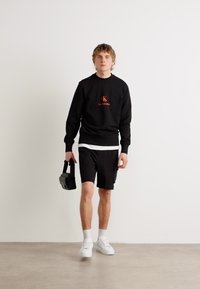 Felpa nera con logo arancione, abbinata a pantaloni neri. Maglietta bianca sotto, sneaker bianche, tiene una piccola borsa nera.
