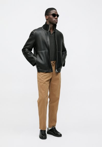 Black leather zip-up jacket with two front pockets, paired with beige trousers and black sneakers. Model wearing sunglasses.