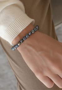 Mustad musta magevee pärlide käevõru, millel on ebakorrapärased vormid ja sinise ning rohelise iridesentsiga, mis puhkab heleda nahatooni randmel.