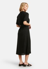 Schwarzes Maxikleid mit kurzen Ärmeln, taillierter Taille und seitlichem Schlitz, aus leichtem Stoff für eine lockere Passform. Getragen mit schwarzen Blockabsatzsandalen.
