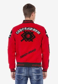 Rote Bomberjacke mit schwarzen bestickten Aufnähern, gerippten Bündchen und Kragen. Enthält den Schriftzug "DENIM BRAND" und geometrische Akzente. Sichtbare Jeans aus Denim.