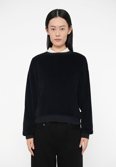 Sudadera de forro polar azul marino con mangas largas, cuello redondo y puños y dobladillo acanalados. Pantalones negros debajo, diseño minimalista.