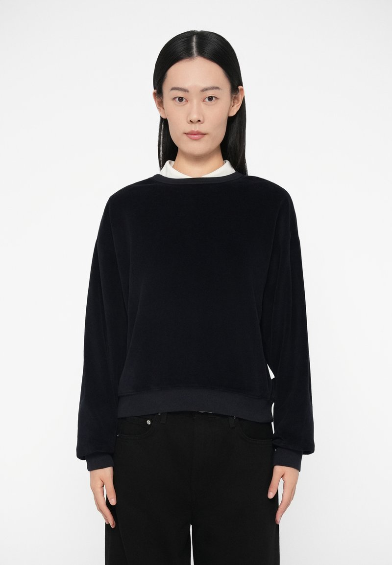 Φούτερ από φλίς σε ναυτικό μπλε, με μακριά μανίκι�α, λαιμόκοψη τύπου crew και ριμπ μανσέτες και τελείωμα. Φοράει μαύρο παντελόνι από κάτω, μινιμαλιστικός σχεδιασμός.