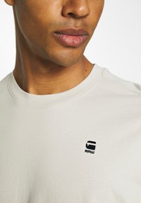 Beige bomull T-skjorte med rund hals og en liten svart logo "RAW" på venstre side nær kragen. Glatt tekstur.
