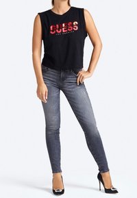 Mujer vistiendo una blusa sin mangas negra de "GUESS Los Angeles", jeans skinny grises descoloridos y tacones altos negros con una hebilla decorativa.