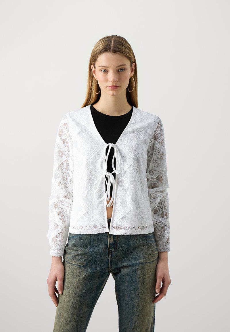Cardigan en dentelle blanche à manches longues, avec un col en V et une fermeture à nouer à l'avant. Porté sur un haut noir et associé à un jean.