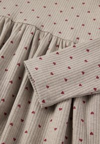 Ribbede beige stofkjole med små røde hjerte-mønstre. Har en samlet talje og lange ærmer. Teksturen er blød og behagelig.