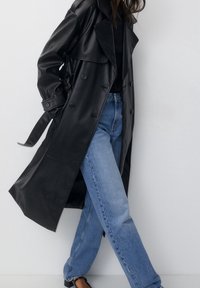 Femme portant un trench-coat en cuir noir, un haut noir, un jean bleu et des chaussures noires, se tenant avec les jambes croisées contre un mur blanc.