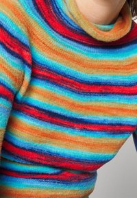 Personne portant un pull en laine doux à rayures multicolores avec des bandes horizontales rouges, bleues, orange et beige, et un col roulé ample.