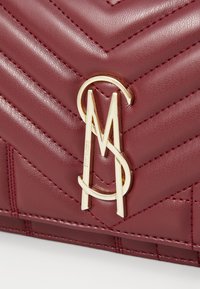 Burgundi nahkakäsilaukku, jossa on chevron-nauhakuva, kullanvärinen logo ja vastakkaiset ommeltu yksityiskohta.