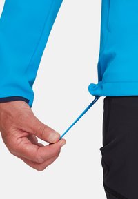 Blaue Jacke mit glatter Textur, die mit einer Hand am Reißverschlusszug angepasst wird. Im unteren Teil des Bildes sind schwarze Hosen sichtbar.