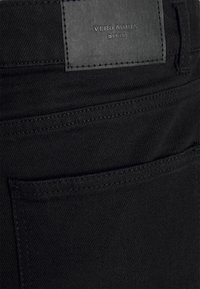 Jeans de ganga preta com um acabamento texturizado, cintura baixa e um bolso traseiro com um patch do logótipo Vero Moda. Detalhes de costura subtis visíveis.
