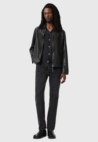 Black leather jacket with zipper, buttoned black shirt underneath, dark jeans, and black boots. Model stands against a light gray background.