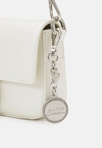 Weißer, texturierter Handtasche mit einem Magnetverschluss, ausgestattet mit einer silbernen Kette und einem runden Anhänger mit "ALDO Collection" und einem Strassrahmen.