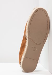 Tan ruskindssko med flad sål, der har en vævet tekstur ved bunden og et præget "GUESS 7M" mærkat. Vinklet hældesign.