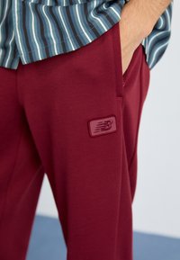 Maroon sweatpants with a smooth texture, featuring a stitched New Balance logo patch and side pockets. Styled with a striped shirt above.