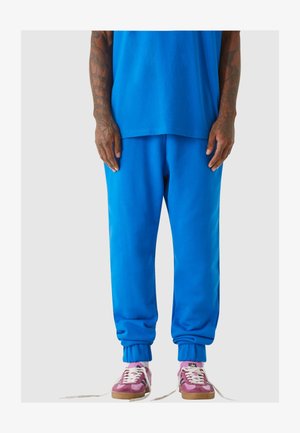 Žiarivo modré tepláky s rebrovanými manžetami a uvoľneným strihom, spárované s ružovými a hnedými teniskami s bielymi šnúrkami a textúrovaným dizajnom.