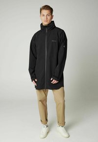 Čierna nepremokavá bunda s vysokým golierom, predným zipsom a nastaviteľnými manžetami. V kombinácii s svetlo béžovými nohavicami a bielymi topánkami.