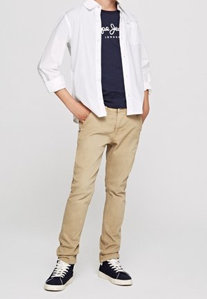 Lekka, biała koszula z guzikami noszona na granatowej koszulce, w zestawieniu z khaki spodniami oraz granatowymi trampkami z białymi podeszwami i sznurówkami.