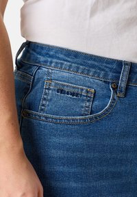 Blue denim jeans with a classic cut, featuring a back pocket with stitching detail and a subtle brand label. Smooth texture and mid-rise waist.