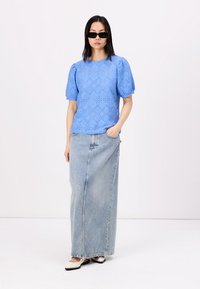 Blouse brodée bleu clair avec des manches bouffantes, associée à une longue jupe en denim. Modèle portant des lunettes de soleil et des chaussures noires et blanches.
