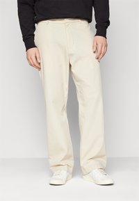 Beige bomullbukser med rett passform, med forkantfold og sidelommer, brukt sammen med hvite joggesko. Minimalistisk design.