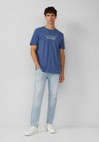 Niebieska koszulka z krókim rękawem z białym napisem "S.Oliver Elevated Casual." W zestawie z jasnoniebieskimi dżinsami i białymi sneakersami.