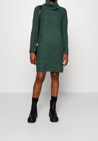 Grön stickad polotröja klänning med långa ärmar, rak skärning, ribbad textur och sidoklackar. Matchas med svarta grova stövlar och strumpor.