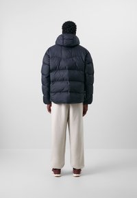 Svart vadderad jacka med luva och kviltat mönster. Bärs med krämfärgade byxor och bruna skor. Minimalistisk stil.